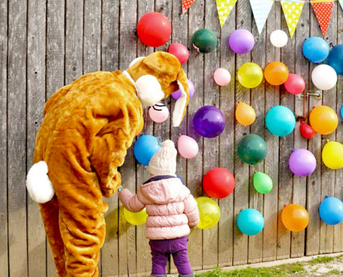 Lebensgroßer Osterhase mit Kind in rosa Jacke und Mütze,sowie viele bunte Luftballons an der Holzwand