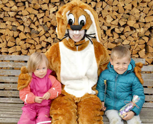 Großer Osterhase umarmt zwei Kinder, dahinter ist eine Holzschar