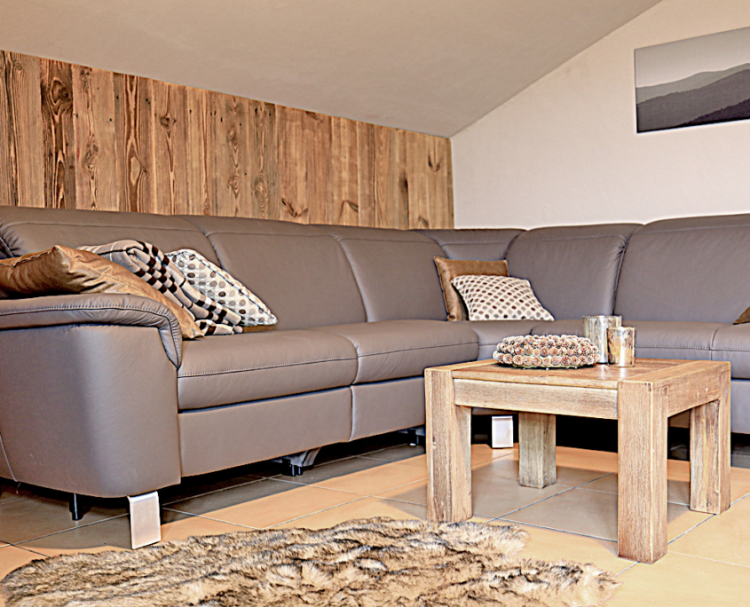 Wohnbereich mit Holzwand, einen Tisch, Couch mit Polster und Dece, sowie ein Fell auf dem Boden